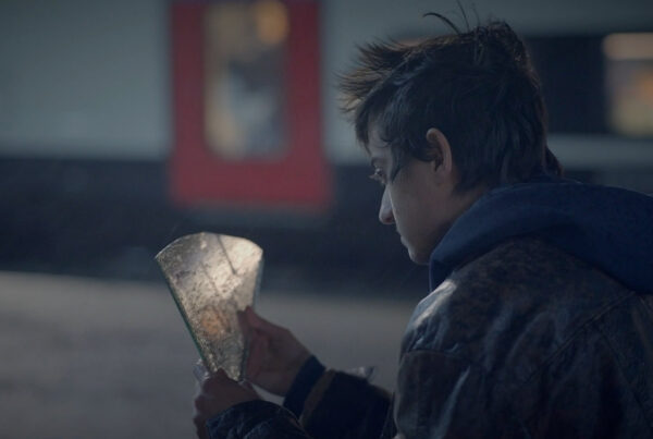 A young man sits on the floor at a train platform, staring at a shard of mirror with the word 'future' written on it. From the Mossbeard song - Failing at it