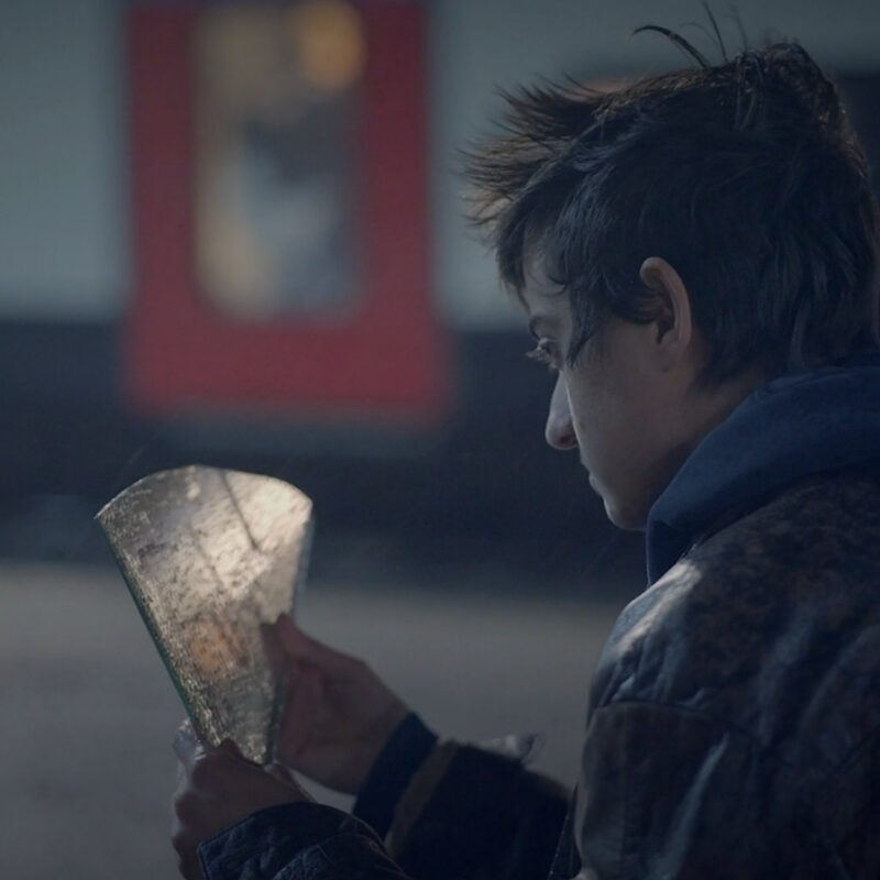 A young man sits on the floor at a train platform, staring at a shard of mirror with the word 'future' written on it. From the Mossbeard song - Failing at it