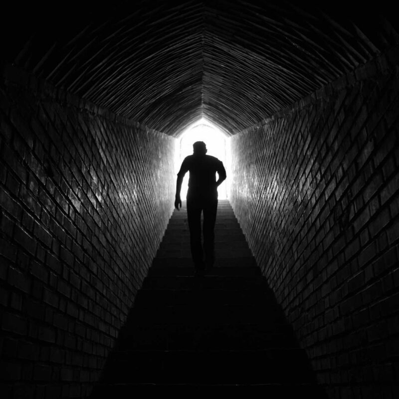 A silhouette of a strong young man walk up a stairway leading to the top of a tunnel and into the light.