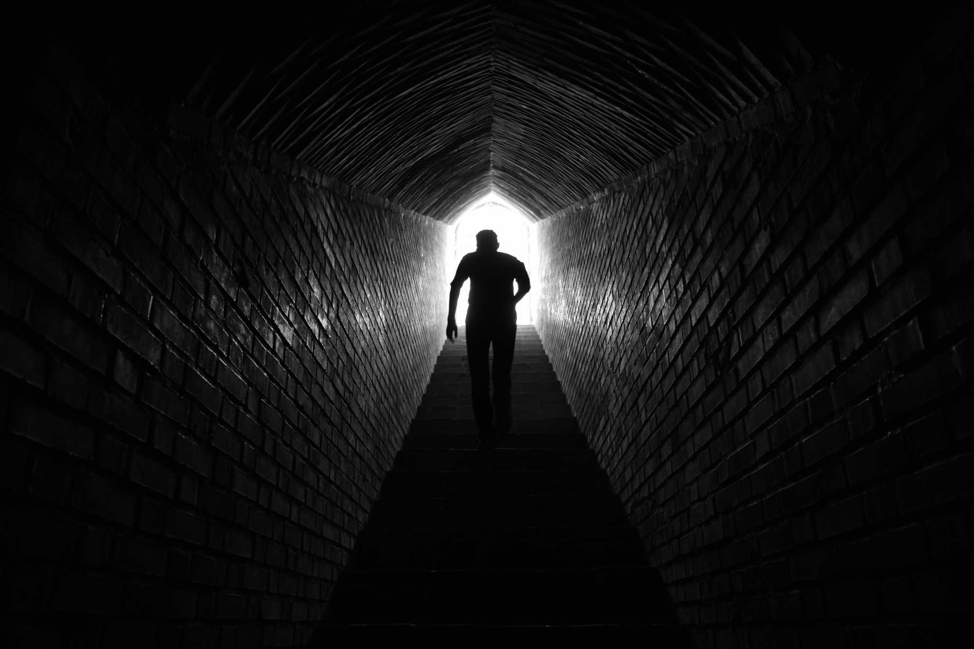 A silhouette of a strong young man walk up a stairway leading to the top of a tunnel and into the light.