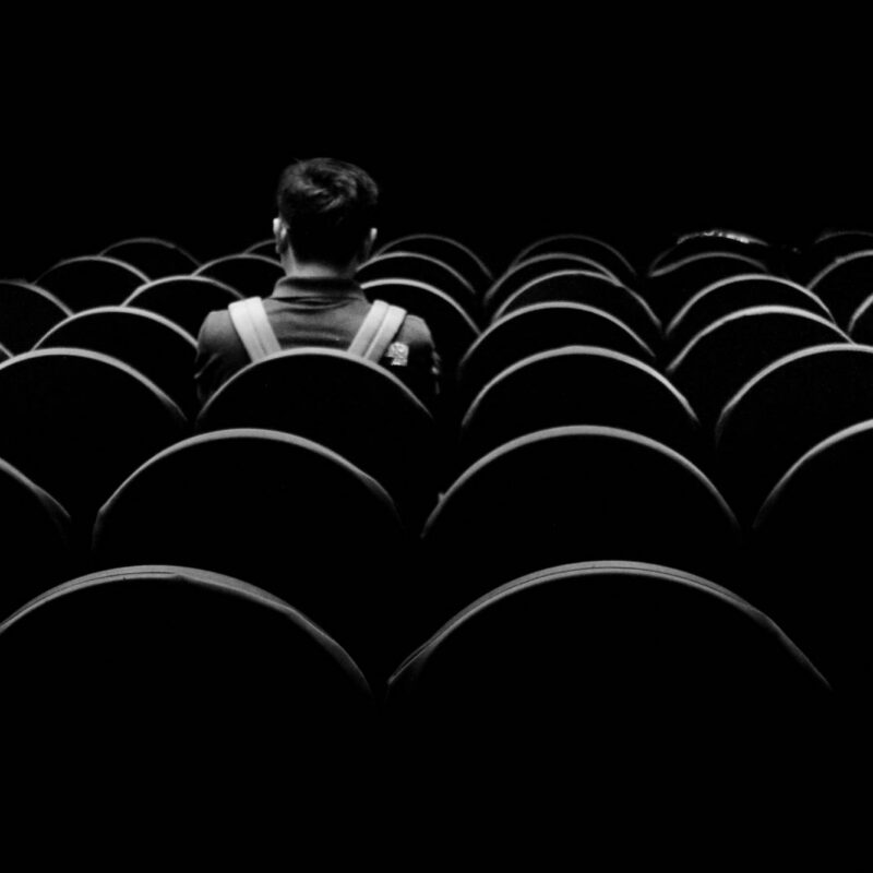 A rear view of a young man sitting alone in an auditorium. Black and white moody.