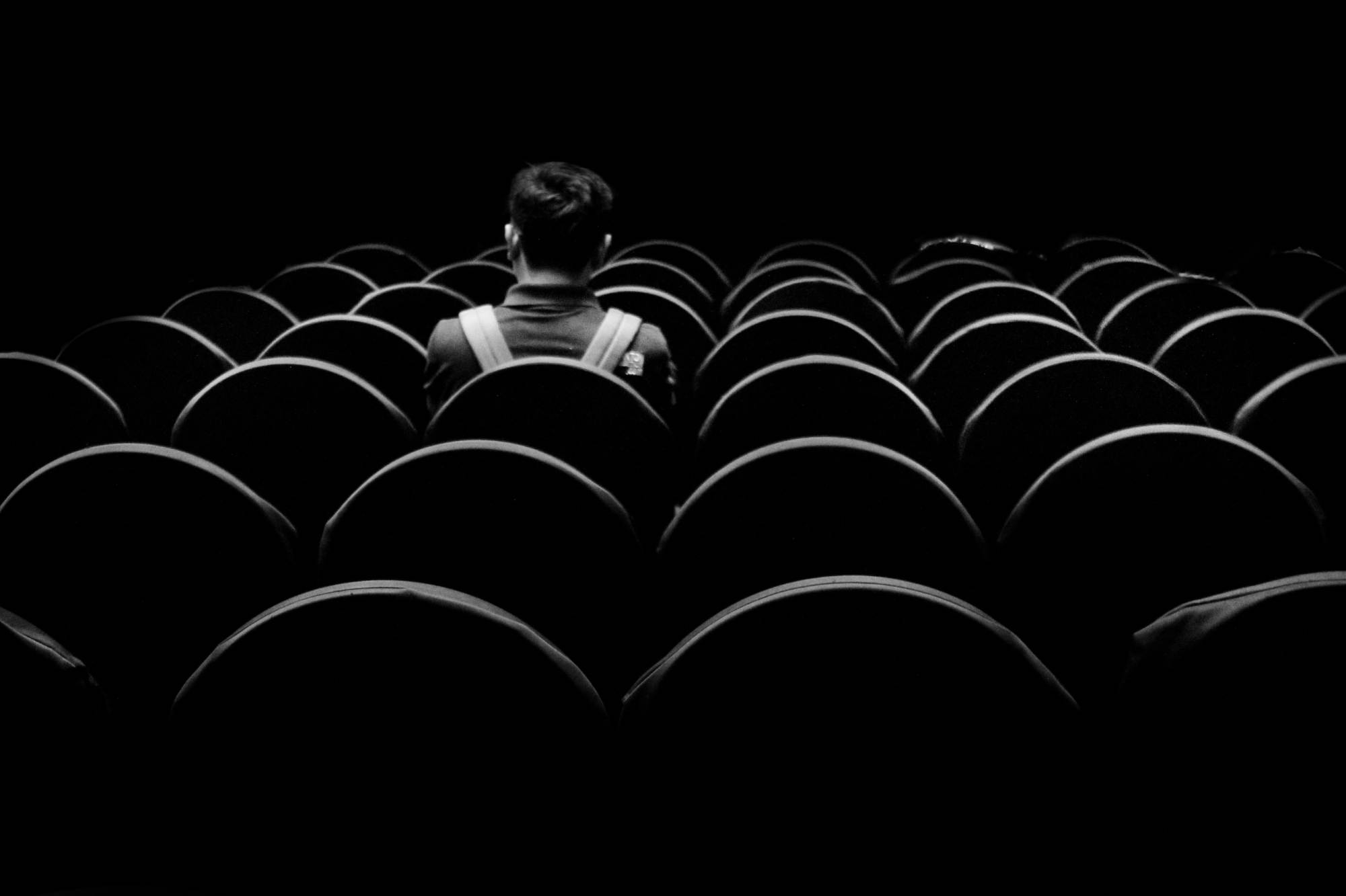 A rear view of a young man sitting alone in an auditorium. Black and white moody.
