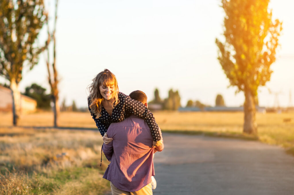 Mossbeard song - You're mine - Rear view of young cheerful man carrying his beautiful girlfriend on shoulder while walking by the country road, the girl looks around and laughs.