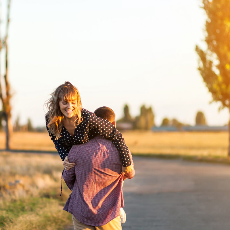 Mossbeard song - You're mine - Rear view of young cheerful man carrying his beautiful girlfriend on shoulder while walking by the country road, the girl looks around and laughs.